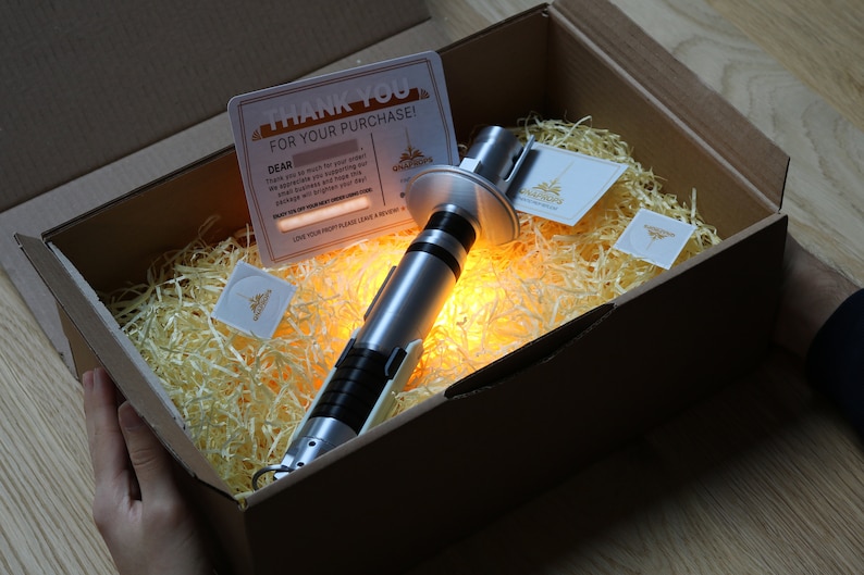 May include: A silver lightsaber prop in a cardboard box with shredded paper. The box is open and the lightsaber is lit up with a warm yellow glow. The box also contains a thank you card and a small business card.