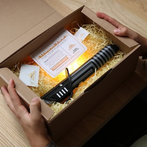 May include: A black and silver lightsaber toy in a brown cardboard box with shredded paper. The box has a thank you note for the purchase.