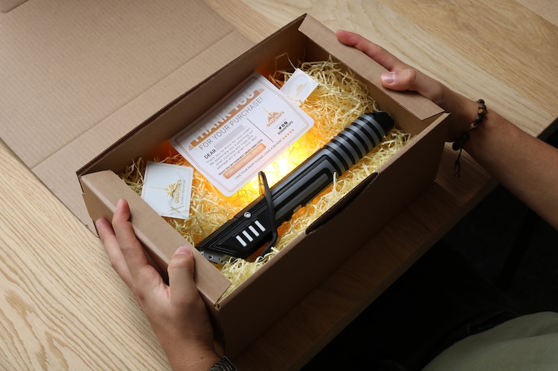 May include: A black and silver lightsaber toy in a brown cardboard box with shredded paper. The box has a thank you note for the purchase.