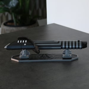 May include: A black and silver lightsaber prop on a stand with a Mandalorian symbol. The lightsaber is on a black and silver stand with a Mandalorian symbol.