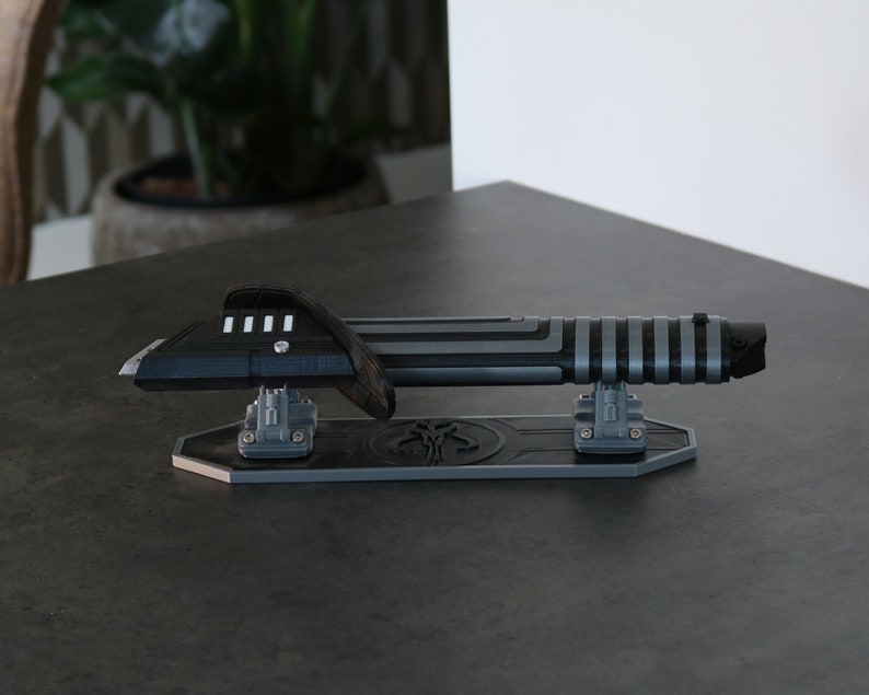 May include: A black and silver lightsaber prop on a stand with a Mandalorian symbol. The lightsaber is on a black and silver stand with a Mandalorian symbol.