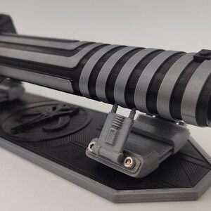 May include: A black and silver lightsaber with a textured grip is displayed on a black and gray stand. The stand has a Mandalorian symbol on it.