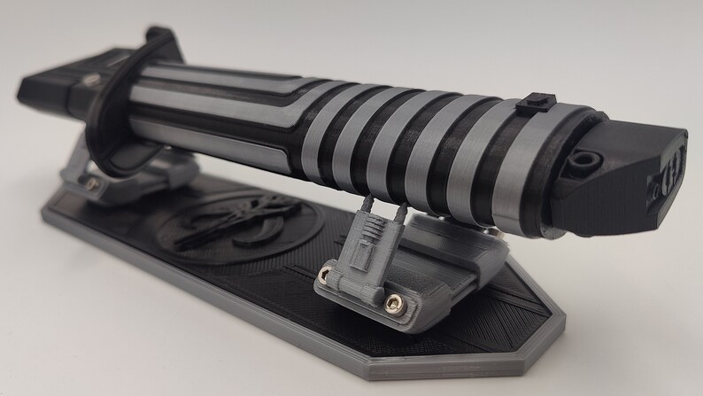 May include: A black and silver lightsaber with a textured grip is displayed on a black and gray stand. The stand has a Mandalorian symbol on it.
