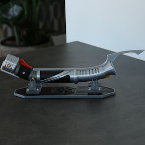 Puede incluir: Un sable de luz plateado y negro con un mango amarillo y rojo, exhibido en un soporte negro con un diseño circular.