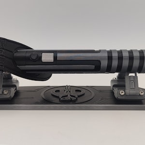 May include: A black and gray lightsaber-shaped USB drive on a gray stand with a black logo. The drive has a black handle and a gray blade with black stripes.