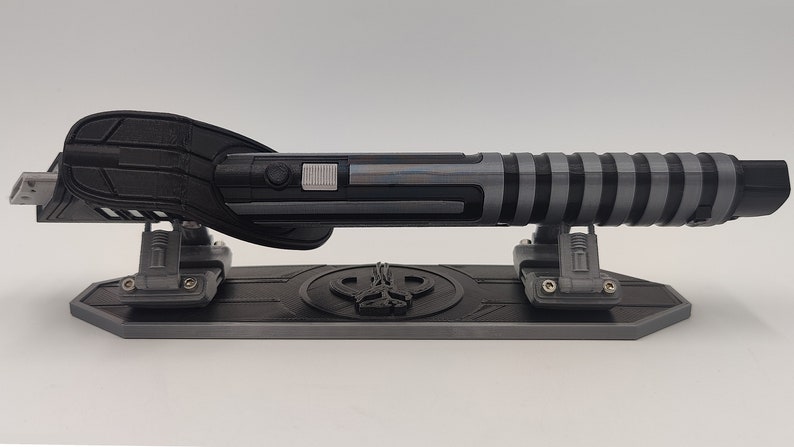 May include: A black and gray lightsaber-shaped USB drive on a gray stand with a black logo. The drive has a black handle and a gray blade with black stripes.