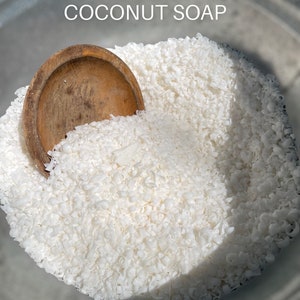 May include: A close-up of a bowl filled with white, hand-grated coconut flakes. The flakes are piled high and a wooden spoon is partially submerged in the flakes. The text "HANDMADE HAND GRATED COCONUT SOAP" is displayed above the bowl.