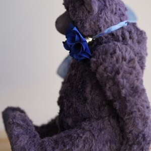 May include: A plush purple teddy bear sitting on a wooden surface. The bear is wearing a blue ribbon around its neck with a blue flower accent.