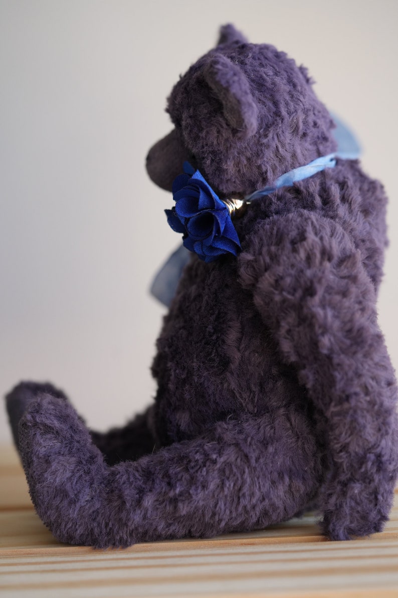 May include: A plush purple teddy bear sitting on a wooden surface. The bear is wearing a blue ribbon around its neck with a blue flower accent.