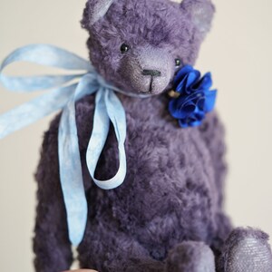 May include: A plush purple teddy bear with a blue flower tied around its neck. The bear is sitting on a hand.
