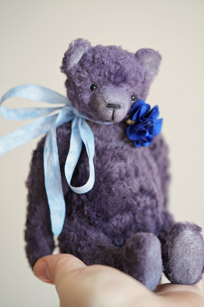 May include: A plush purple teddy bear with a blue flower tied around its neck. The bear is sitting on a hand.