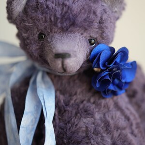 May include: A purple teddy bear with a blue flower tied around its neck. The bear has a blue ribbon tied around its neck.