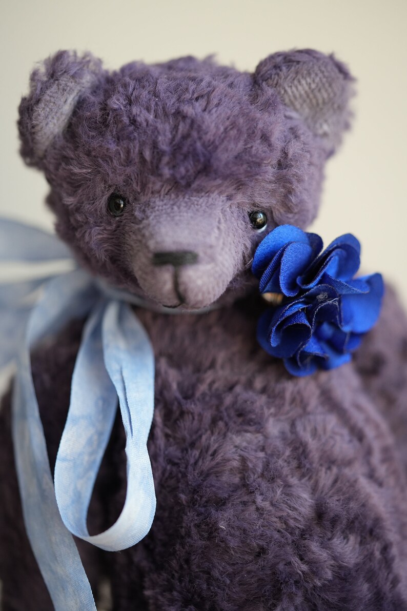 May include: A purple teddy bear with a blue flower tied around its neck. The bear has a blue ribbon tied around its neck.