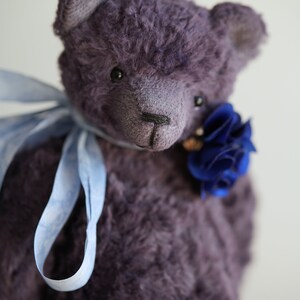 May include: A plush purple teddy bear with a blue flower on its chest and a light blue ribbon around its neck.