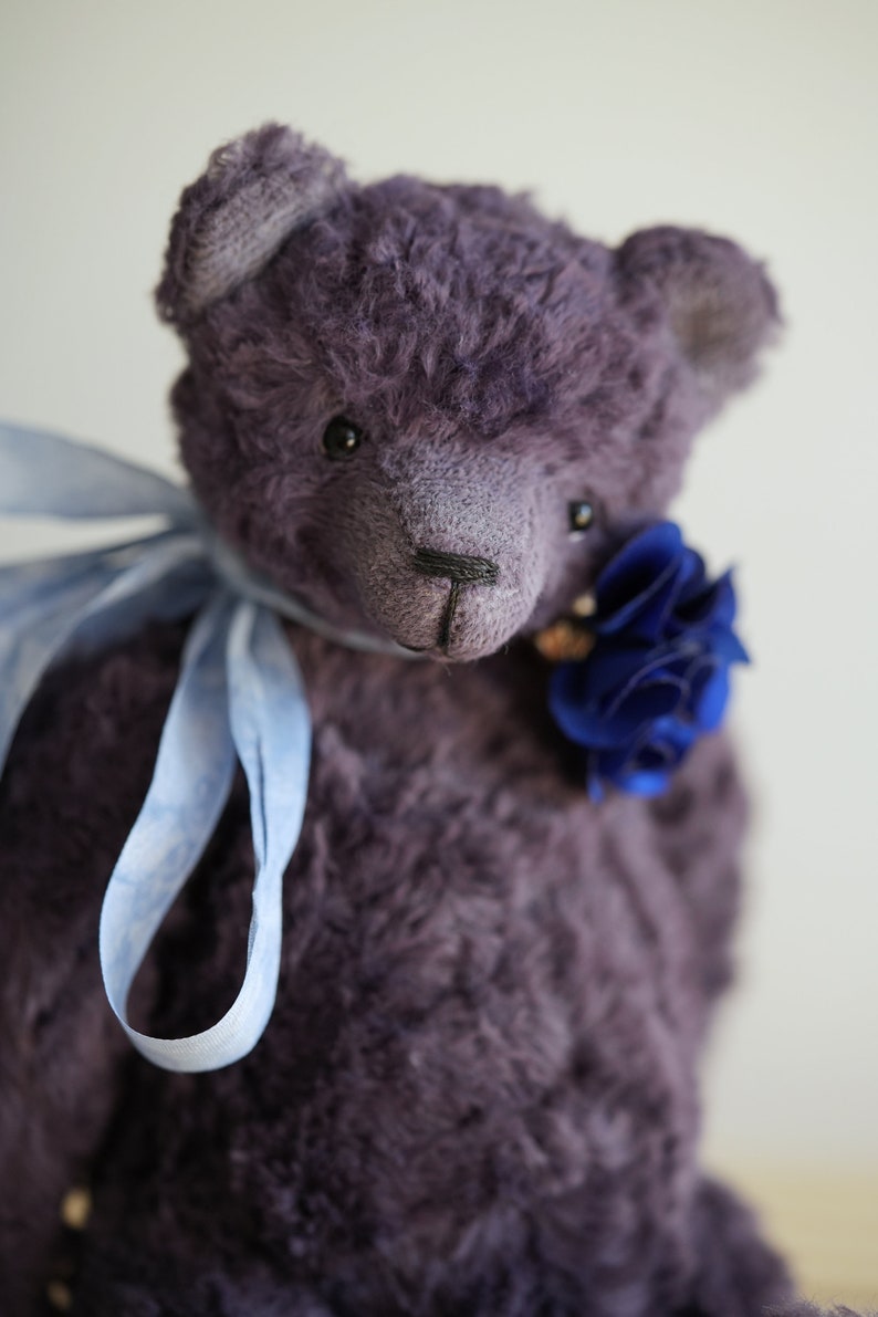 May include: A plush purple teddy bear with a blue flower on its chest and a light blue ribbon around its neck.
