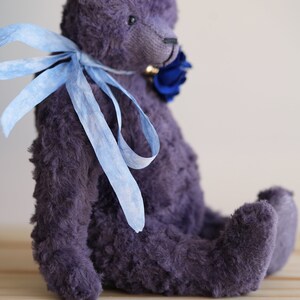 May include: A plush purple teddy bear with a blue ribbon around its neck. The bear is sitting on a wooden surface.