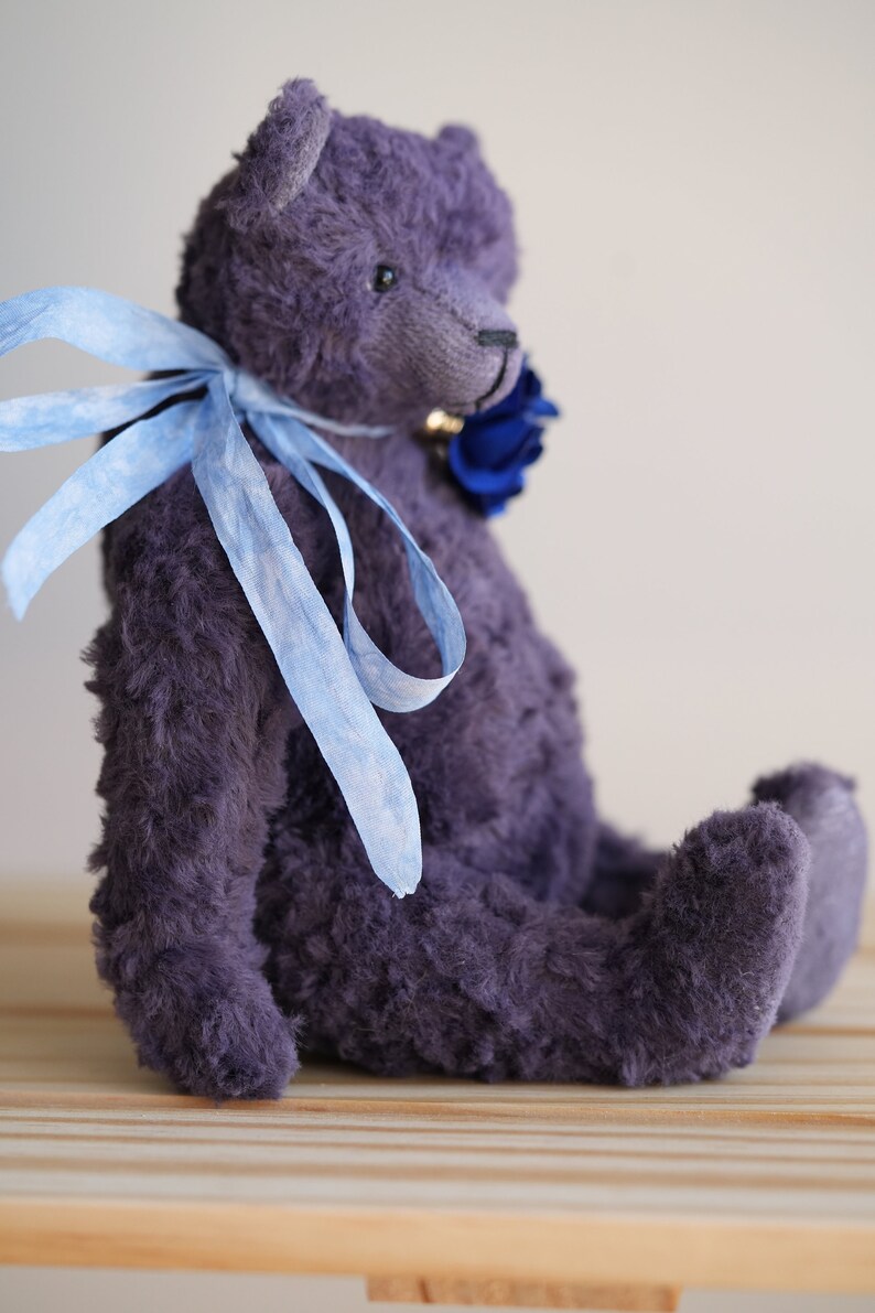 May include: A plush purple teddy bear with a blue ribbon around its neck. The bear is sitting on a wooden surface.