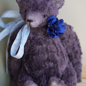 May include: A plush purple teddy bear with a blue flower on its chest. The bear is sitting on a wooden surface.