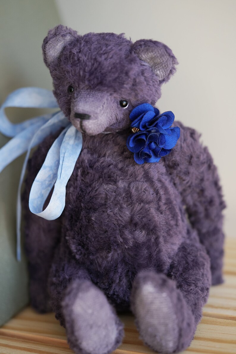 May include: A plush purple teddy bear with a blue flower on its chest. The bear is sitting on a wooden surface.