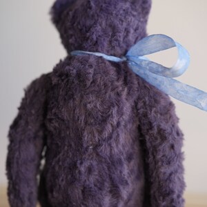 May include: A plush purple teddy bear with a blue ribbon tied around its neck. The bear is sitting on a wooden surface.