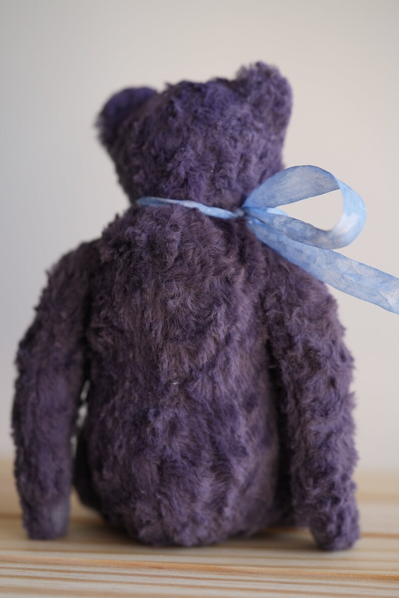 May include: A plush purple teddy bear with a blue ribbon tied around its neck. The bear is sitting on a wooden surface.