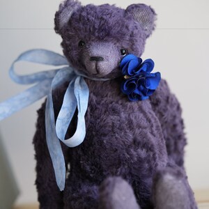 May include: A plush purple teddy bear with a blue flower on its chest and a blue ribbon around its neck. The bear is sitting on a wooden surface.
