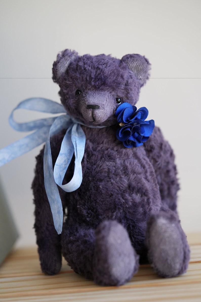 May include: A plush purple teddy bear with a blue flower on its chest and a blue ribbon around its neck. The bear is sitting on a wooden surface.