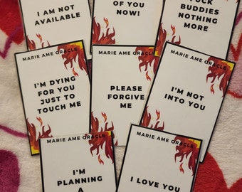 May include: A collection of white oracle cards with black text and a fiery red and orange flame design. The cards feature phrases like "I AM NOT AVAILABLE" and "I LOVE YOU". The brand name "MARIE AME ORACLE" is printed at the top of each card.