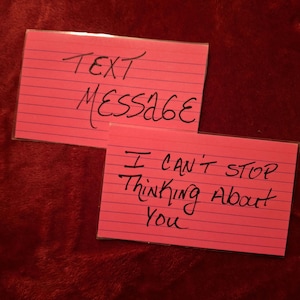 May include: Two pink index cards with handwritten text. One card reads "TEXT MESSAGE" and the other reads "I CAN'T STOP THINKING ABOUT YOU". The cards are on a red, textured surface.