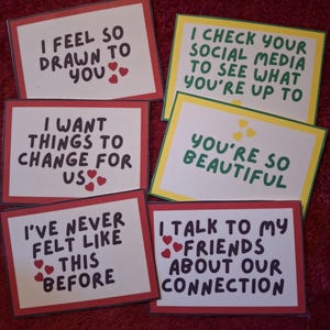 May include: A collection of six handmade cards with handwritten messages and heart illustrations. The cards have red, yellow, and white borders, and the text includes phrases like "I feel so drawn to you" and "You're so beautiful."