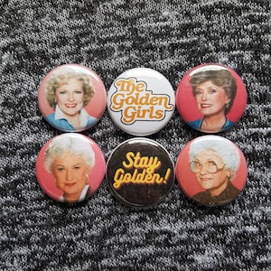 Puede incluir: Seis botones redondos con personajes del programa de televisión "The Golden Girls". Los botones muestran el texto "The Golden Girls" y "Stay Golden!" en letras amarillas sobre un fondo negro. Los botones también muestran imágenes de los cuatro personajes principales: Bea Arthur, Rue McClanahan, Estelle Getty y Betty White.