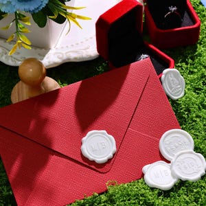 May include: A textured red envelope sealed with a white wax seal featuring the letters M|B. Nearby are three additional white wax seals with various monograms, a red velvet ring box containing a ring, and a wooden stamp handle.