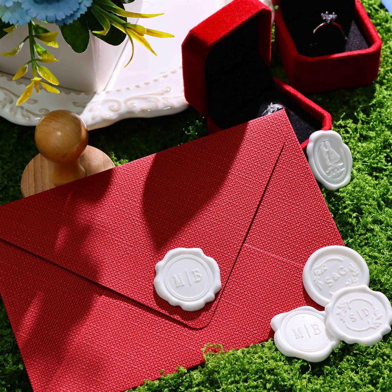 May include: A textured red envelope sealed with a white wax seal featuring the letters M|B. Nearby are three additional white wax seals with various monograms, a red velvet ring box containing a ring, and a wooden stamp handle.