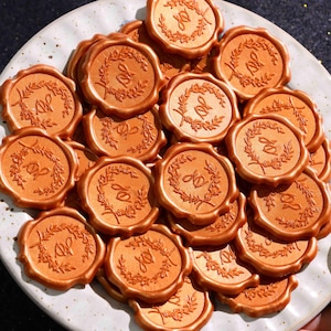 May include: A collection of round, metallic copper-colored wax seals piled on a white plate. Each seal features a raised embossed design of a circular laurel wreath surrounding the intertwined cursive letters JD in the center of the wax surface.