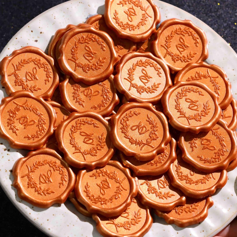 May include: A collection of round, metallic copper-colored wax seals piled on a white plate. Each seal features a raised embossed design of a circular laurel wreath surrounding the intertwined cursive letters JD in the center of the wax surface.