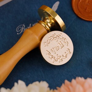 May include: A gold-toned wax seal stamp with a wooden handle. The stamp features a wreath design with the initials "V & M" in the center.