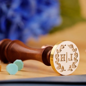 May include: A gold-toned wax seal stamp with a wreath design and the initials "H|J" engraved on it. The stamp is attached to a wooden handle.