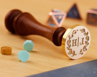 Puede incluir: Un sello de madera con una cabeza de latón que presenta un diseño de corona de laurel y las iniciales "H|J".