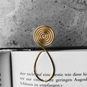 May include: A gold-colored metal bookmark with a spiral design at the top and a teardrop shape below. The bookmark is placed in an open book with German text. The background is a textured gray surface.