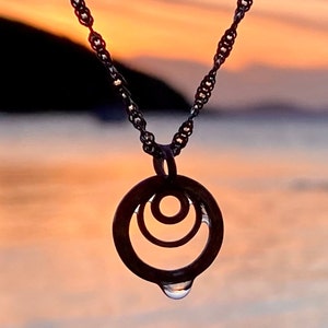 May include: A silver necklace with a pendant featuring three concentric circles, the outer circle has a teardrop hanging from the bottom. The necklace is photographed against a blurred background of a sunset over water.