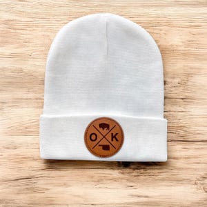 May include: White beanie with a brown leather patch featuring the letters "OK" with a buffalo and the outline of Oklahoma.