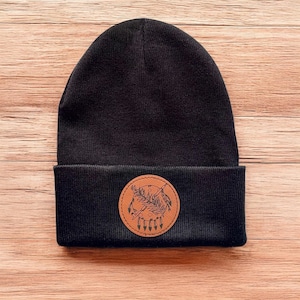 May include: Black beanie with a brown leather patch featuring a Native American design. The design includes a feather, a spear, and a circle with arrows.