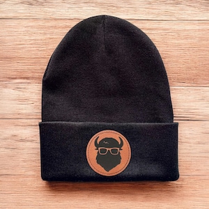 May include: Black beanie with a brown leather patch featuring a silhouette of a buffalo wearing glasses.