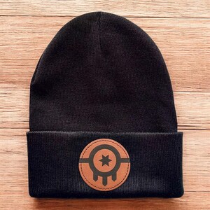 May include: Black knit beanie with a brown leather patch featuring a stylized star and teardrop design.