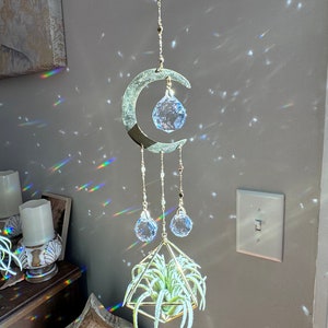 May include: A celestial-themed hanging air plant holder. The design features a crescent moon, crystal orbs, and a geometric gold-colored frame holding a green air plant. The piece is suspended by a chain and creates a play of light.