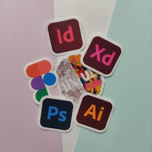 Può includere: Una collezione di adesivi colorati con i loghi di Adobe Creative Suite, tra cui Photoshop, Illustrator, InDesign, After Effects e XD. Gli adesivi sono disposti su uno sfondo rosa chiaro, bianco e azzurro.