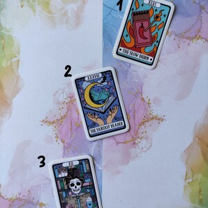 K&ouml;nnte beinhalten: Drei Tarotkarten-Aufkleber mit bunten Illustrationen und Text. Der erste Aufkleber ist blau mit einer roten Chilischote und Streichh&ouml;lzern und dem Text "XLVI THE SLOW BURN". Der zweite Aufkleber ist lila mit einem Halbmond, Bergen und H&auml;nden, die ein Buch halten, und dem Text "XXXVII THE FANTASY READER". Der dritte Aufkleber ist lila mit einem Sch&auml;del, der eine Krone tr&auml;gt und ein Buch h&auml;lt, und dem Text "XII THE LIBRARIAN".