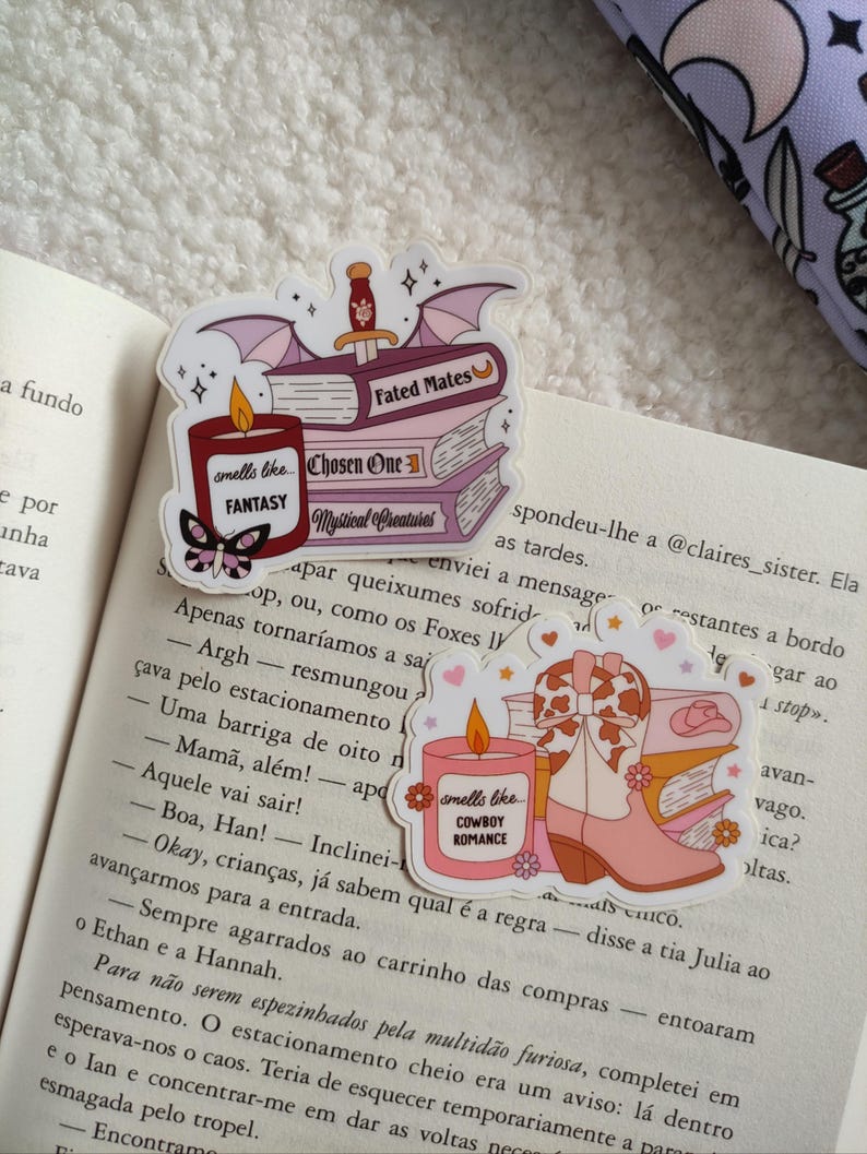 May include: Three pink and purple sticker designs featuring stacks of books with a candle on top. The candles are labeled "smells like... FANTASY" and "smells like... COWBOY ROMANCE". The books are labeled "Fated Mates", "Chosen One", and "Mystical Creatures".