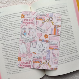 Pode incluir: Um marcador de livros rosa com um padrão floral e temático de livros, com frases como "I'm All Booked" e "Bookish". O marcador de livros está colocado num livro aberto, com texto visível nas páginas.