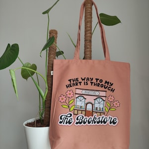 Może przedstawiać: Różowa torba z płótna z napisem "The way to my heart is through The Bookstore" i kreskówkową ilustracją księgarni z kwiatami.